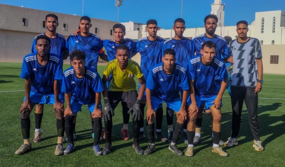 inbound3955271721915703340 - جامعة سبها | Sebha University Group of young soccer players in blue jerseys pose for a team photo on a green field with buildings and a clear blue sky in the background, front row kneeling.