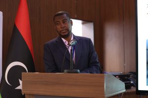 Man in a dark suit speaking at a wooden podium with a microphone in a conference room, flag in the background