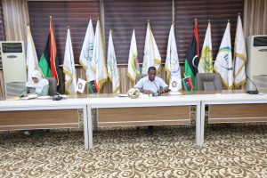 inbound4836711866385244713 - جامعة سبها | Sebha University Two officials sit at a long conference table signing documents, with many flags behind them in a formal meeting room.