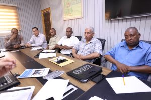 Six men sit around a conference table in a meeting room, papers and folders scattered on the table as they look toward the camera.