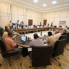 A formal meeting in a large conference room with delegates seated around a U-shaped table, Libyan flags displayed, several laptops and documents on the table.