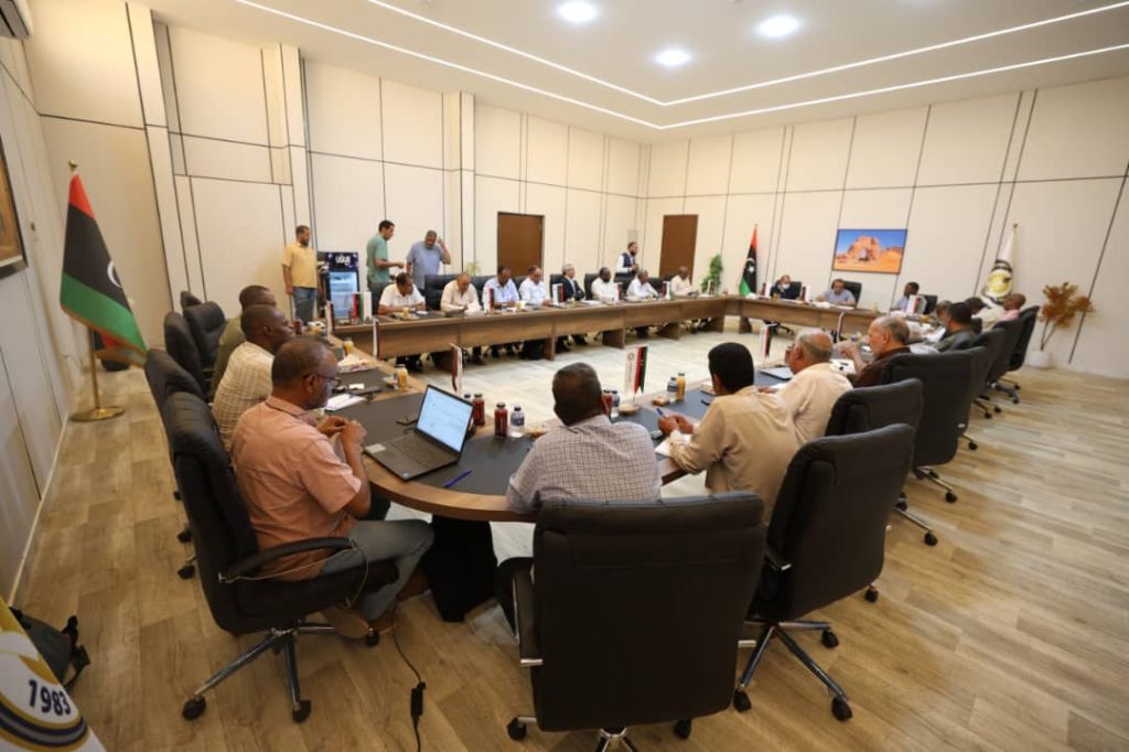 inbound5067218647295939110 - جامعة سبها | Sebha University A formal meeting in a large conference room with delegates seated around a U-shaped table, Libyan flags displayed, several laptops and documents on the table.