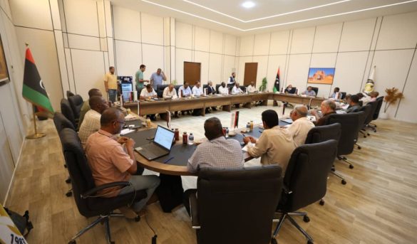 inbound5067218647295939110 - جامعة سبها | Sebha University A formal meeting in a large conference room with delegates seated around a U-shaped table, Libyan flags displayed, several laptops and documents on the table.