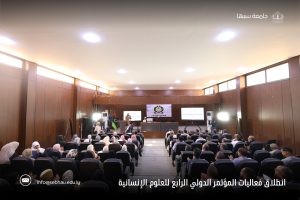 inbound5106752767038852715 - جامعة سبها | Sebha University Audience seated in a wood-paneled conference hall facing a stage with banners and screens showing presentations.