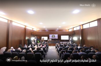 Audience seated in a wood-paneled conference hall facing a stage with banners and screens showing presentations.