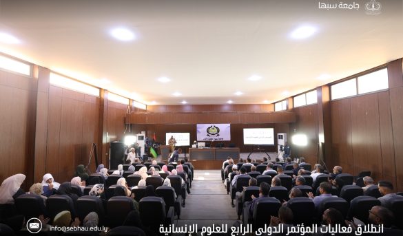 inbound5106752767038852715 - جامعة سبها | Sebha University Audience seated in a wood-paneled conference hall facing a stage with banners and screens showing presentations.