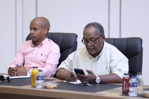 inbound5253442824691454685 - جامعة سبها | Sebha University Two men sit at a conference table in an office; one writes notes while the other looks at a smartphone during a meeting.