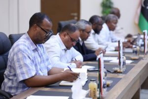 inbound5828845095264052641 - جامعة سبها | Sebha University Group of professionals seated at a long conference table, taking notes during a formal meeting with flags and bottles on the table.