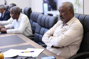 inbound6204889866059049331 - جامعة سبها | Sebha University Man in a beige shirt sits with arms crossed at a conference table, papers and a pen in foreground, other attendees blurred in the background.