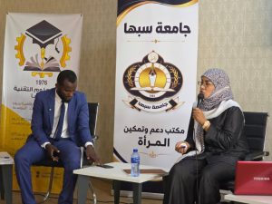 Panel discussion scene: a man in a blue suit and a woman in a patterned hijab speak on stage, with Arabic banners behind them.