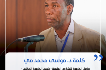 Male speaker in a beige suit delivering a formal talk at a conference, with a microphone in front and a blue lanyard visible.