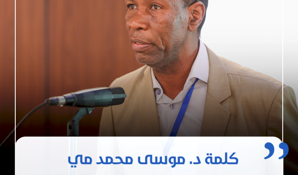 inbound6254940944879704446 - جامعة سبها | Sebha University Male speaker in a beige suit delivering a formal talk at a conference, with a microphone in front and a blue lanyard visible.