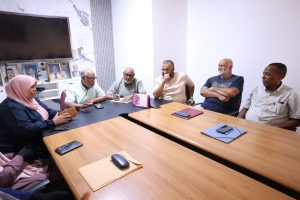 inbound6277397376736346490 - جامعة سبها | Sebha University Six adults sit around a wooden conference table in a bright meeting room; a woman in a pink hijab is on the left and five men across the table, one taking notes.