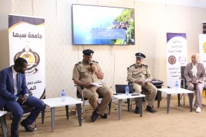 Panel discussion with uniformed officials seated in a conference room, banners in Arabic on either side and a large screen behind them.
