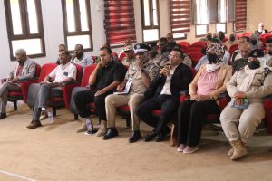 Audience in a conference hall with red chairs; mix of uniformed police officers and civilians wearing masks and hijabs, seated attentively in front row.