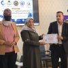 During a certificate presentation: a man in a suit hands a framed certificate to a woman in a headscarf, with a second woman nearby and a uniformed officer in the background.