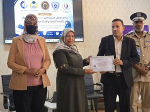 During a certificate presentation: a man in a suit hands a framed certificate to a woman in a headscarf, with a second woman nearby and a uniformed officer in the background.