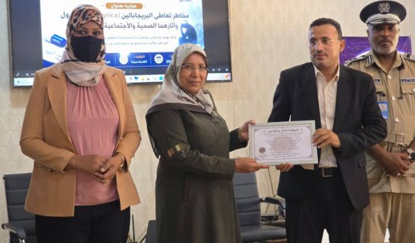 inbound7094141747565374477 - جامعة سبها | Sebha University During a certificate presentation: a man in a suit hands a framed certificate to a woman in a headscarf, with a second woman nearby and a uniformed officer in the background.