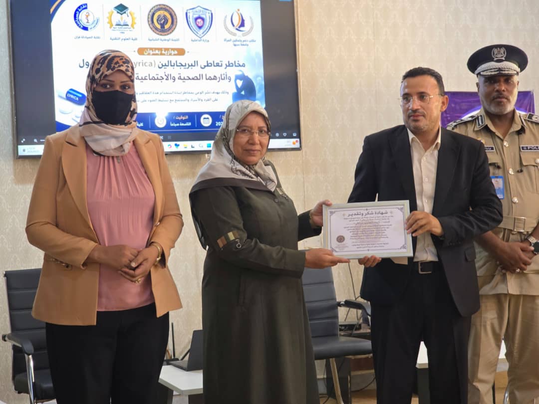 During a certificate presentation: a man in a suit hands a framed certificate to a woman in a headscarf, with a second woman nearby and a uniformed officer in the background.