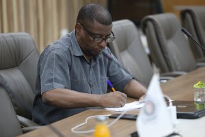 inbound7370345672115915288 - جامعة سبها | Sebha University Man sitting at a conference table, writing on papers, wearing glasses and a blue shirt; microphones and empty chairs visible in the background.