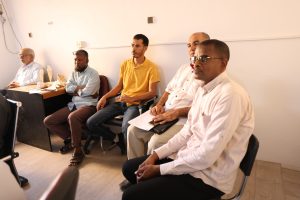 inbound7374796502168525725 - جامعة سبها | Sebha University Five men sit in a row in a meeting room, listening attentively, with papers on a table nearby.