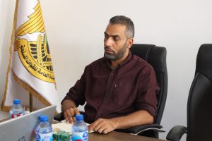 Man in maroon shirt seated at a conference table, looking to his left, with a flag featuring a yellow emblem in the background.