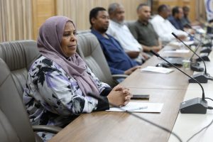 inbound7469380215795474142 - جامعة سبها | Sebha University Front-row woman in a lavender hijab sits at a conference table with notebook and pen, others in background attending meeting.