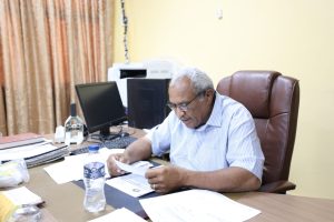 Older man with glasses sits at a cluttered desk in an office, reading documents with computer monitors and a printer in the background.