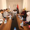 Group of people in a formal meeting around a long conference table, with flags in the background and laptops, water bottles, and notebooks on the table.