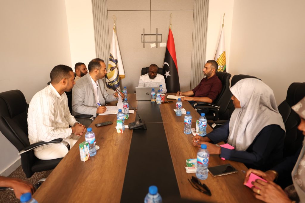 inbound7671426338962303933 - جامعة سبها | Sebha University Group of people in a formal meeting around a long conference table, with flags in the background and laptops, water bottles, and notebooks on the table.