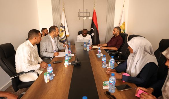 inbound7671426338962303933 - جامعة سبها | Sebha University Group of people in a formal meeting around a long conference table, with flags in the background and laptops, water bottles, and notebooks on the table.