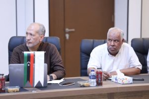 inbound7814128226370248810 - جامعة سبها | Sebha University Two men sit at a conference table in a meeting room, with a flag display, laptop, and drinks on the table between them.