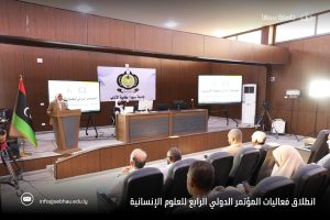 inbound8055523448488916655 - جامعة سبها | Sebha University Speaker at podium in a conference hall with Libyan flag, banner of Sebha University, and panel table on stage; audience seated.