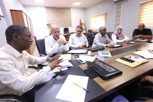 Six professionals sit around a long conference table filled with papers, notebooks, and phones during a meeting in a bright room.