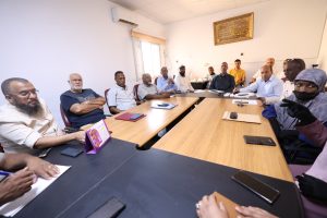 inbound8177238664200558937 - جامعة سبها | Sebha University A diverse group sits around a long conference table in a meeting room, some with notebooks and phones, engaged in a discussion.