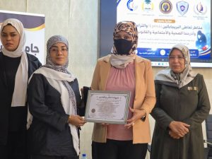 Four women in hijabs stand together at a formal event; the center woman in a peach blazer holds a framed certificate.