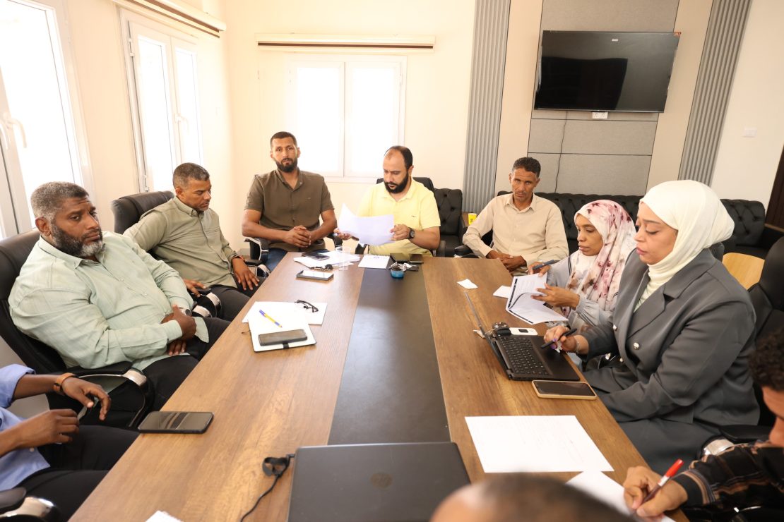 Group of adults seated around a conference table in a bright meeting room, reviewing documents and using laptops.
