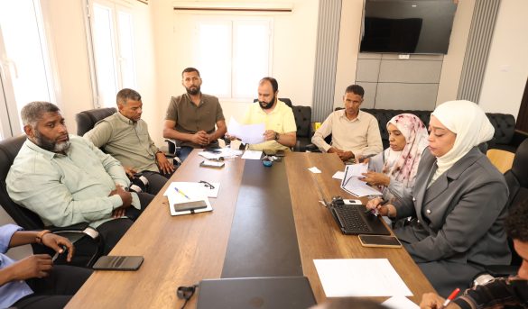 inbound8206219400520692768 - جامعة سبها | Sebha University Group of adults seated around a conference table in a bright meeting room, reviewing documents and using laptops.