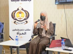 Woman in hijab and black mask speaks into a microphone during a panel discussion, with a banner featuring an emblem and Arabic text behind her and a red laptop on the table nearby.
