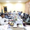 Officials sit around a long conference table in a yellow-walled room, reviewing documents and chatting; Libyan flag in the corner nearby.