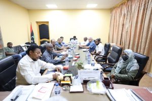 Officials sit around a long conference table in a yellow-walled room, reviewing documents and chatting; Libyan flag in the corner nearby.