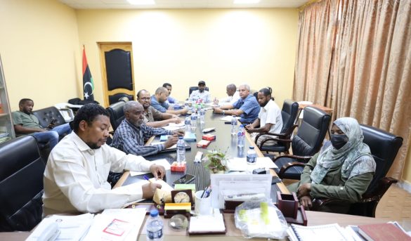 inbound8390770898795824447 - جامعة سبها | Sebha University Officials sit around a long conference table in a yellow-walled room, reviewing documents and chatting; Libyan flag in the corner nearby.