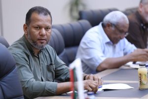 inbound8489477054051779895 - جامعة سبها | Sebha University Man in a green shirt sits at a conference table, facing the camera, with papers and a flag on the desk and a juice bottle nearby.