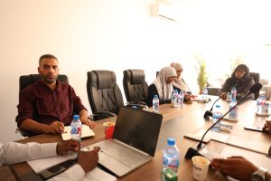 Five people sit around a conference table with laptops, notebooks, and bottled water in a bright meeting room.