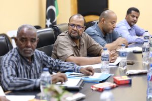 Four men sit at a conference table in a meeting, with papers, notebooks and bottled water on the table.