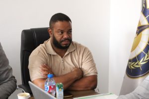 Man with a beard seated at a desk in an office, wearing a light brown polo, arms crossed, with a water bottle and flag in the background.