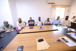 inbound878462027987058997 - جامعة سبها | Sebha University Eight men seated along a large conference table in a bright meeting room, some with papers and phones in front of them, appearing engaged in discussion.