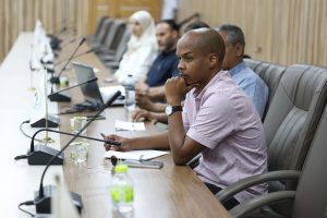 inbound902029818828779772 - جامعة سبها | Sebha University Man in a pink shirt sits at a conference table with his hand on his chin, others with laptops and microphones in the background.
