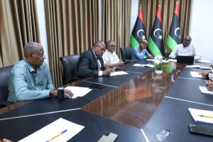Group of men in a formal meeting around a wooden conference table with Libyan flags in the background