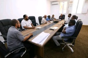 Group of men in business casual sit around a long conference table in a meeting room on a workday (informative scene).